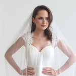 A woman in a white wedding dress with a veil stands confidently with her hands on her hips, gazing directly at the camera.