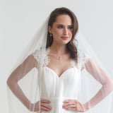 A woman in a white wedding dress with a veil stands confidently with her hands on her hips, gazing directly at the camera.