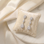 A pair of elegant diamond earrings rests on a white velvet cushion, which is placed on a marble surface.