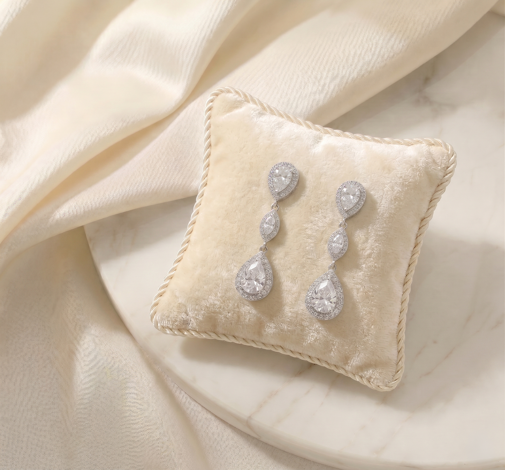 A pair of elegant diamond earrings rests on a white velvet cushion, which is placed on a marble surface.