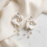 A pair of elegant white earrings with delicate flower-shaped designs and dangling pearls, resting on a white fabric surface.