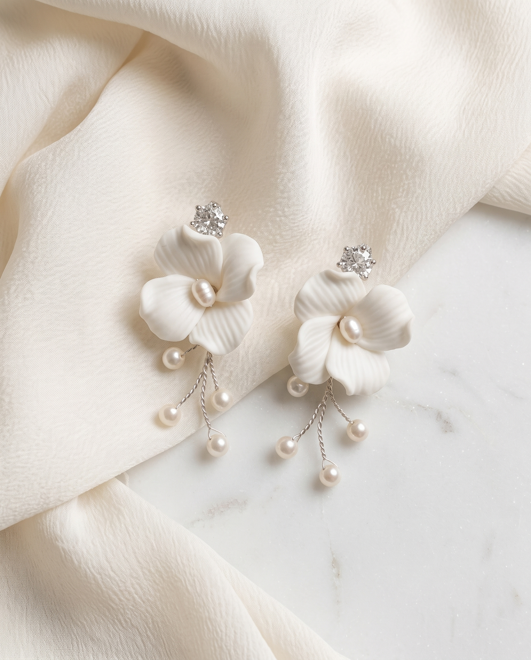 A pair of elegant white earrings with delicate flower-shaped designs and dangling pearls, resting on a white fabric surface.