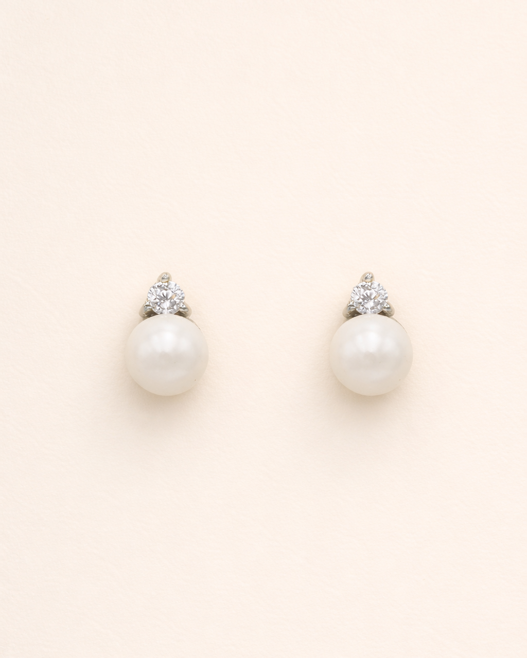 A pair of pearl earrings with a diamond accent, set against a light beige background.