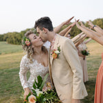 A bride and groom are embracing and kissing in a field, surrounded by their wedding party who are holding bouquets and flowers.
