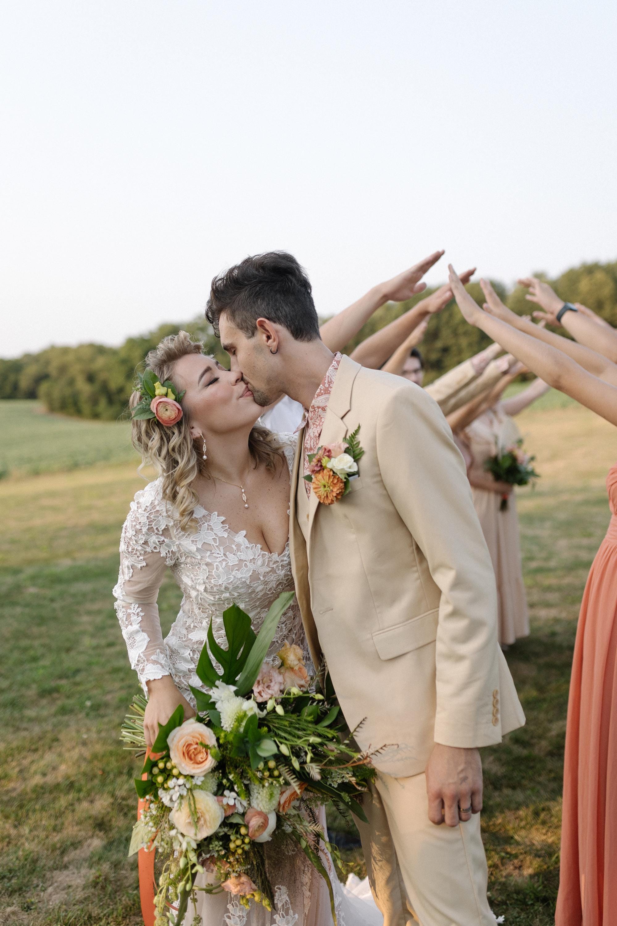 A bride and groom are embracing and kissing in a field, surrounded by their wedding party who are holding bouquets and flowers.